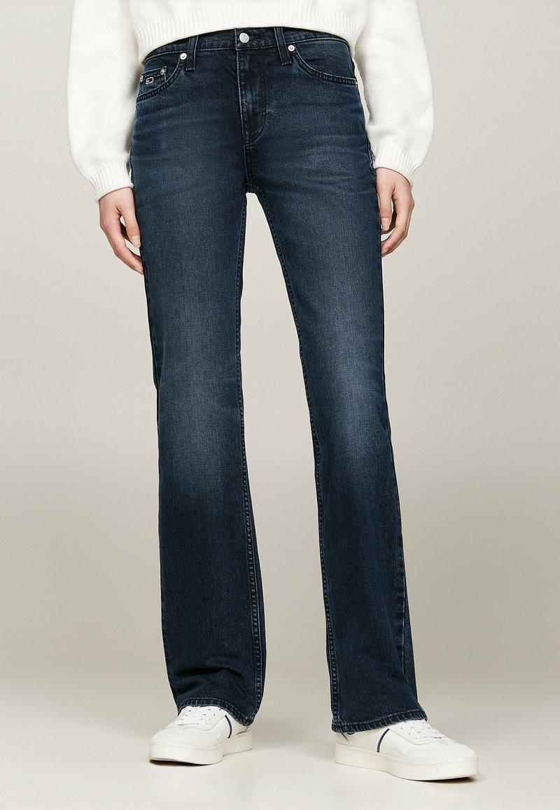Jeans acampanados de un azul oscuro, confeccionados en denim, con una cintura de altura media, diseño de cinco bolsillos y un ligero desvanecimiento en las piernas. Combinados con zapatillas blancas.