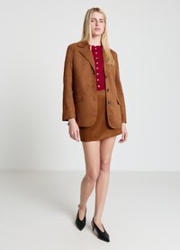 Brown suede blazer with notch lapels, single-breasted buttons, and two front pockets, paired with a fitted maroon top and skirt. Black heels.