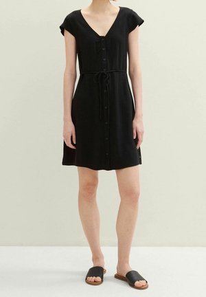 Kobieta ubrana w czarną sukienkę z krótkim rękawem zapinaną na guziki z paskiem w talii oraz czarne sandały na płaskim obcasie, stojąca na jasnym, jednolitym tle.