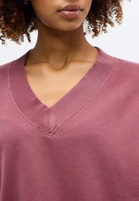 Γυναίκα με σγουρά μαλλιά φορώντας πουλόβερ με V λαιμόκοψη σε μωβ απόχρωση, που δείχνει το ντεκολτέ και το πάνω μέρος του στήθους.