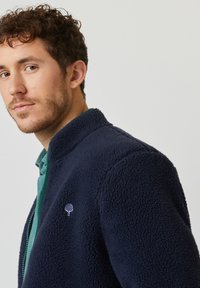 Marineblaue Fleecejacke mit hohem Kragen und Reißverschluss. Mit einem kleinen, gestickten Baum-Logo auf der Brust. Weicher Stoff.