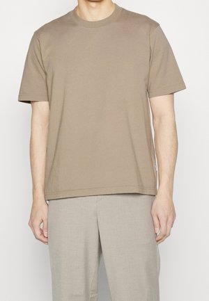 Homme portant un t-shirt marron uni à manches courtes et un pantalon beige clair, debout sur un fond blanc uni.