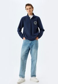 Sudadera con cremallera de color azul marino con un logotipo circular en el lado izquierdo del pecho, combinada con jeans de color azul claro y zapatillas blancas, de pie contra un fondo blanco.