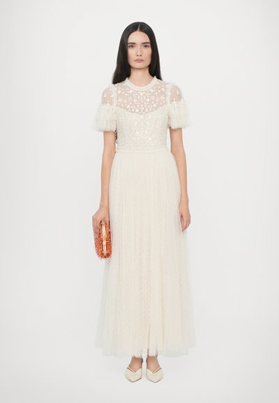 Witte vloerlange jurk van doorschijnende geborduurde stof, korte gerimpelde mouwen en een tailleband. Model houdt een roze parel clutch vast en draagt crèmekleurige schoenen.