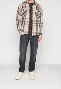 Chemise en flanelle à carreaux pour homme en beige avec des lignes marron et bleues, équipée de deux poches poitrine, associée à un jean sombre et des baskets blanches.