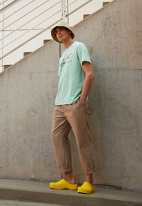 Hellgrünes T-Shirt mit schwarzem Grafikdesign, beigefarbene Cropped-Hosen und leuchtend gelbe Clogs. Der Hintergrund ist aus Beton.