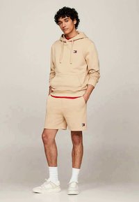 Hanorac bej din bumbac cu șnururi, buzunar tip kangaroo și patch cu logo. Asortat cu pantaloni scurți bej și adidași albi cu detalii texturate.