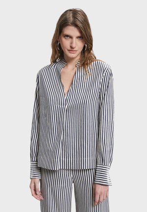 Kobieta w dopasowanej granatowo-białej koszuli i spodniach w pionowe paski z długimi rękawami oraz kolczykami kołami, stojąca na jednolitym tle.