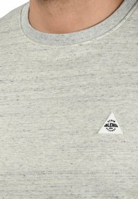 Sudadera gris claro hecha de una tela texturizada, con un cuello redondeado y un pequeño parche de logo triangular blanco en el pecho.