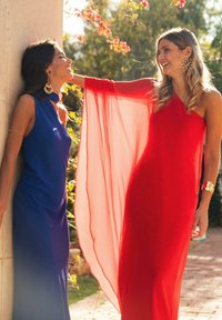 Dos mujeres con vestidos largos: uno azul con un collar choker, el otro rojo con un forro transparente. Ambas llevan pendientes llamativos, situadas al aire libre con acentos florales.