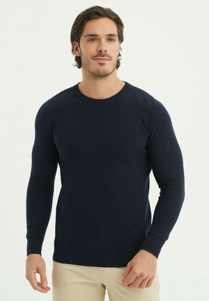 Pull en tricot bleu marine à manches longues et col rond. Il présente des poignets et un ourlet côtelés, ajoutant texture et structure.