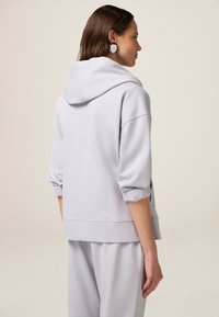Felpa grigia chiara in materiale morbido con vestibilità rilassata, spalle cadute e maniche con polsini. La vista posteriore mostra un design minimalista e i dettagli del cappuccio.