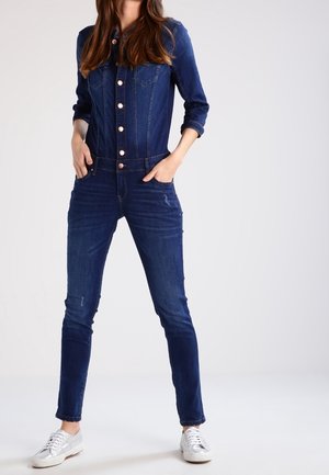 Kvinde iført en tætsiddende mørkeblå denim-jumpsuit med knapper foran, hænderne i lommerne og sølvfarvede sneakers, stående på en hvid baggrund.