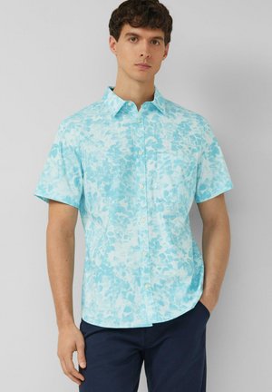 Młody mężczyzna w jasnoniebieskiej kwiecistej koszuli z krótkim rękawem i ciemnoniebieskich spodniach, stoi z jedną ręką w kieszeni.