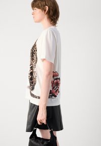 T-shirt crème à manches courtes avec un imprimé léopard et un design floral. Porté avec une jupe en simili cuir noire et un sac à main noir.