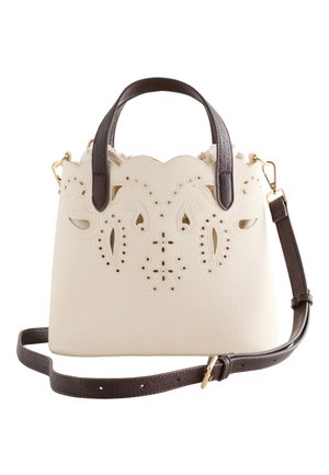 Hand hält cremefarbene Handtasche mit floralem Ausschnittdesign und dunklen Griffen vor schwarzem Kleid-Hintergrund.