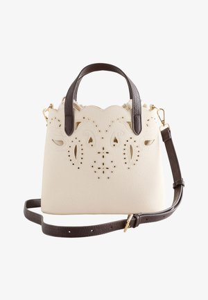 Hand hält cremefarbene Handtasche mit floralem Ausschnittdesign und dunklen Griffen vor schwarzem Kleid-Hintergrund.