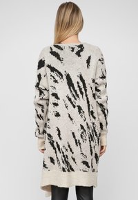 Beżowy sweter kardigan z czarnymi abstrakcyjnymi wzorami. Długie rękawy, zaokrąglony dekolt i dłuższy tylny dół tworzą luźny krój.
