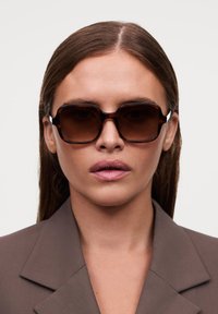 Ochelari de soare supradimensionați, de culoare maro, cu un cadru glossy gros și lentile întunecate, având o textură netedă și accente albe contrastante pe laterale.