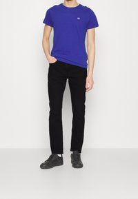 Osoba stojící před bílým pozadím, oblečená v modrém tričku, černých kalhotách, bílých ponožkách a černých teniskách.