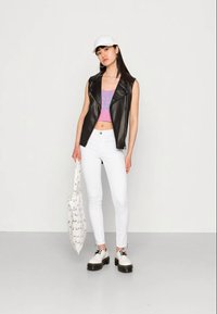 Colete de couro preto sem mangas sobre um top curto rosa, calças skinny brancas e sapatos de plataforma brancos; segurando uma bolsa branca com detalhes em preto.