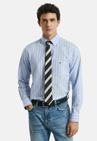 Camicia a righe blu chiaro con colletto button-down, taschino sul lato sinistro, abbinata a una cravatta a righe multicolore e jeans scuri.