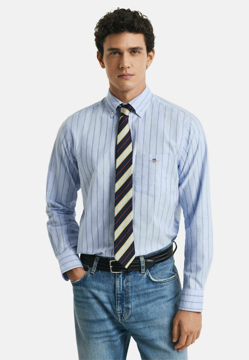 Camicia a righe blu chiaro con colletto button-down, taschino sul lato sinistro, abbinata a una cravatta a righe multicolore e jeans scuri.