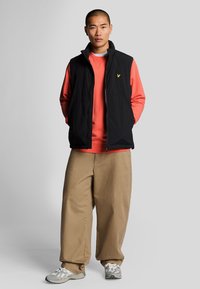 Zwarte mouwloze vest over een rode sweatshirt, kaki broek en grijze sneakers. Bevat een geel logo op het vest. Eenvoudig ontwerp en sportieve fit.