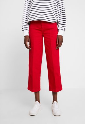 Personne portant un pantalon large rouge raccourci, un haut à rayures blanches et bleu marine à manches longues, et des baskets blanches, se tenant devant un fond uni.