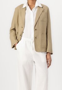 Giacca beige in lino con una vestibilità rilassata, caratterizzata da due tasche frontali, tre bottoni e una camicia bianca col colletto sotto.