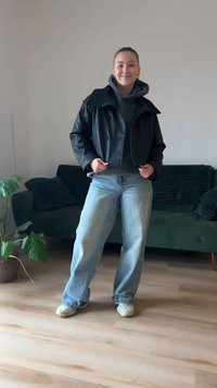 Svart skinnjakke over en grå hettegenser, kombinert med vide lyseblå jeans og hvite joggesko. Innendørs setting med grønn sofa og plante.
