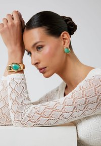 Gioielli in oro con accenti di pietre verdi. Top bianco lavorato a maglia con un motivo a diamante. Superficie liscia e lucidata con forme definite.