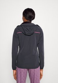 Veste légère noire avec capuche, présentant des accents roses le long des épaules, ainsi que des poignets et un ourlet élastiqués pour une coupe ajustée.