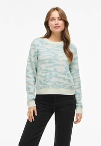 Maglione azzurro chiaro con tessuto morbido e strutturato, con motivo a nuvole e scollo rotondo. Polsini e orlo sono di un azzurro chiaro uniforme. Jeans neri.