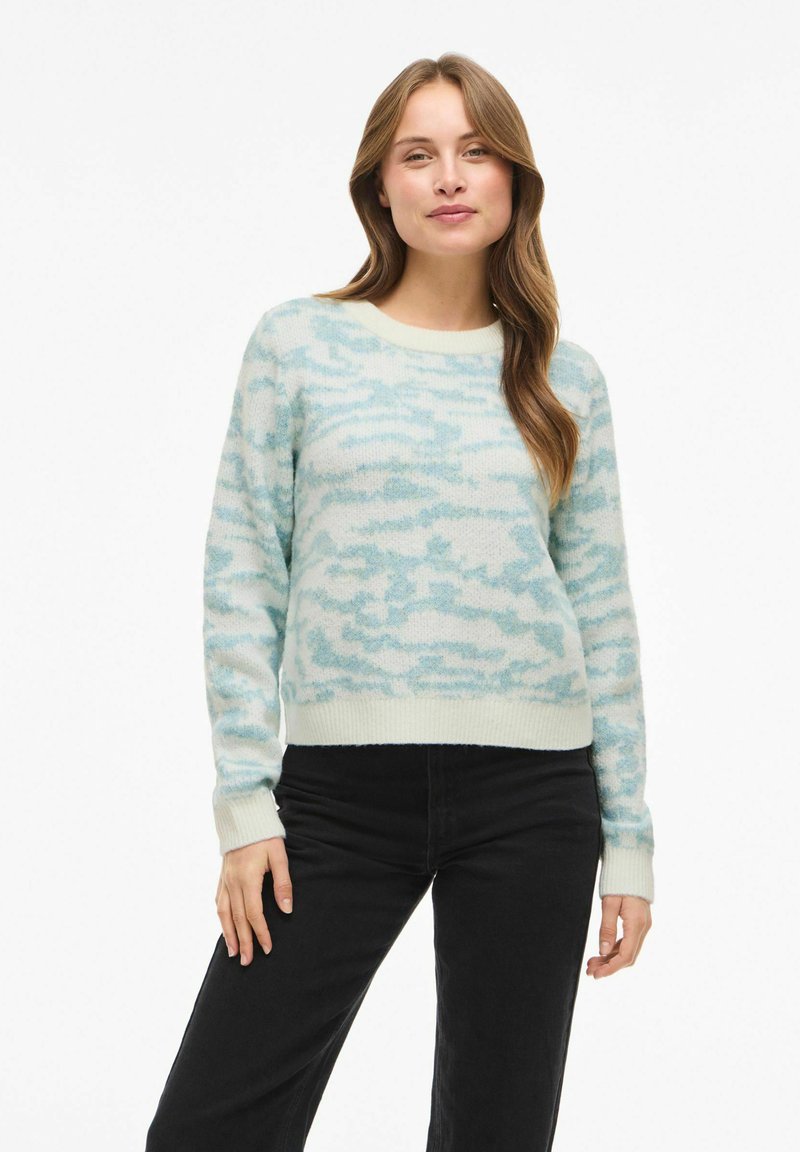 Maglione azzurro chiaro con tessuto morbido e strutturato, con motivo a nuvole e scollo rotondo. Polsini e orlo sono di un azzurro chiaro uniforme. Jeans neri.