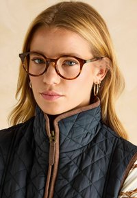 Tortišķu brillēm ar apaļām kārtām, valkātas ar tumši zilu siltinātu vestu ar brūnu apkakli un rāvējslēdzēju. Redzami zelta apli auskari.