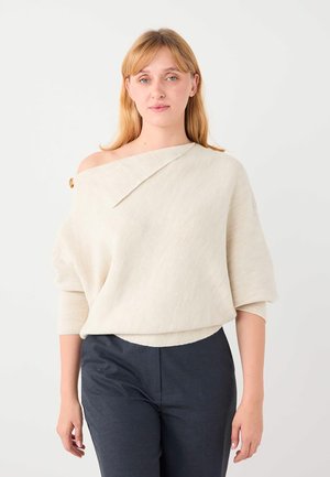 Jonge vrouw met blond haar, gekleed in een off-shoulder crèmewitte trui en donkere broek, staand tegen een effen witte achtergrond.