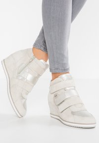 Jasnoszare sneakersy na koturnie z metalicznie srebrnymi paskami i zamkiem błyskawicznym, noszone z dopasowanymi szarymi dżinsami na białym tle.