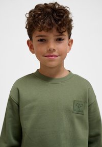 Ung dreng med krøllet brunt hår og fregner iført en olivengrøn sweatshirt med en bi-logo på brystet, smilende blidt.