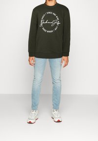 Felpa verde scuro con logo e testo bianchi, polsi a coste, abbinata a jeans skinny blu chiaro e sneakers bianche con dettagli rossi.