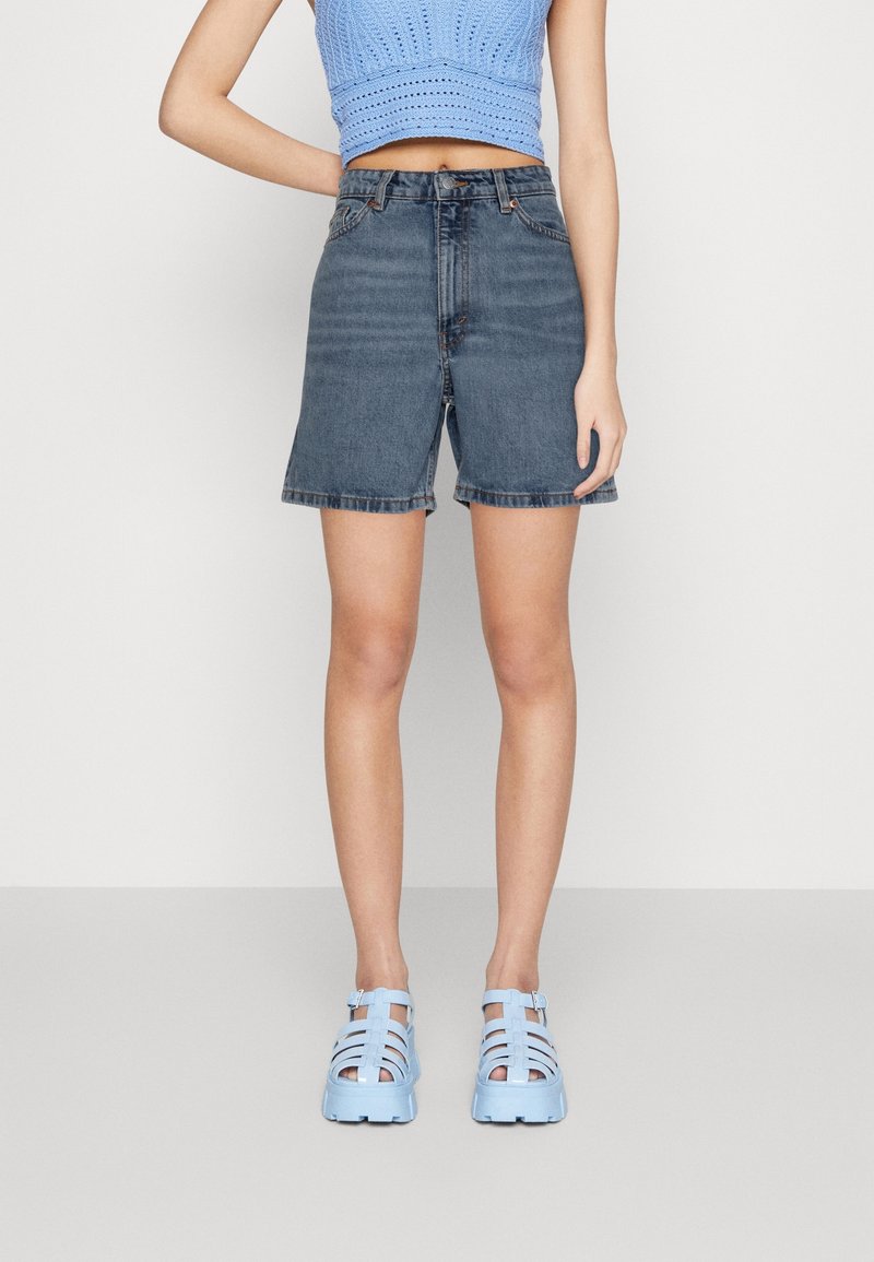Femme portant un crop top bleu tricoté, un short en jean sombre taille haute et des sandales bleu clair fermées, debout devant un fond uni.