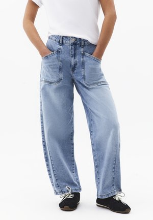 Person trägt hellblaue Jeans mit weitem Bein und großen aufgesetzten Taschen vorne, ein weißes Hemd und schwarze Turnschuhe mit weißen Schnürsenkeln, Hände in den Taschen.