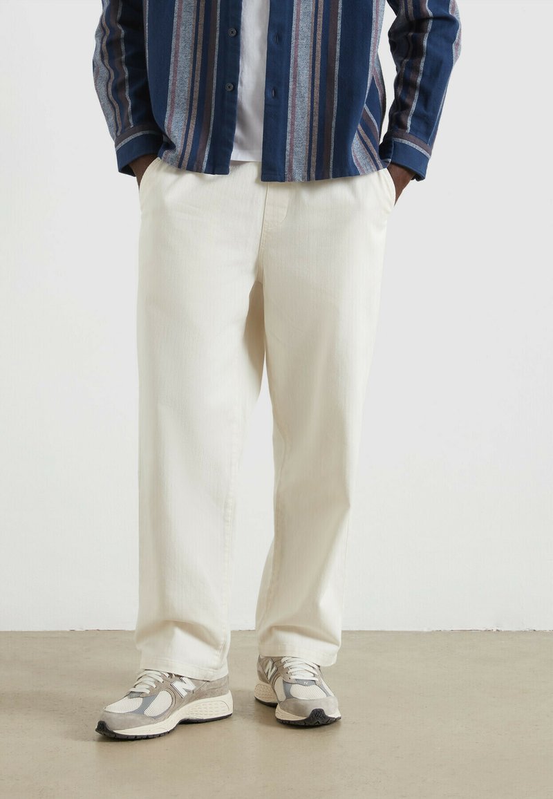 Jasno-kremowe, luźne spodnie wykonane z miękkiego materiału, w zestawieniu z szaro-beżowymi butami sportowymi. Koszula w poziome niebiesko-szare paski.