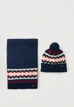 Écharpe tricotée bleu marine et bonnet assorti avec motif Fair Isle rouge, blanc et vert et pompon sur le bonnet, sur fond blanc.