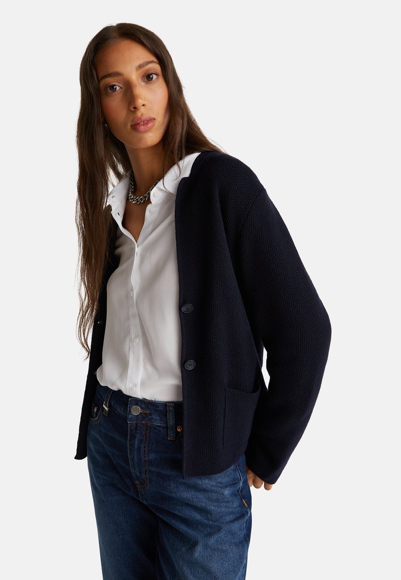 Marineblauer strukturierter Cardigan mit zwei Knöpfen und Fronttaschen, über einem weißen Hemd getragen, kombiniert mit dunkelblauen Jeans.
