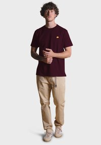 Bordová bavlněná tričko s malým žlutým logem slona, spárované s béžovými kalhotami a béžovými teniskami s jemnými akcenty.