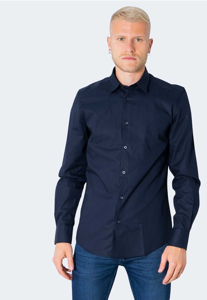 Camicia blu navy a maniche lunghe con bottoni in tessuto liscio, colletto classico e bottoni frontali, abbinata a jeans blu aderenti.