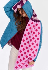 Modrá prešívaná bunda s kapucňou; bordový sveter; ružový šál deka s červenými bodkami prehodený cez ramená, kontrastné textúry a farby.