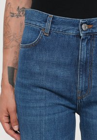 Vaqueros de mezclilla en azul oscuro con un corte clásico. Presenta un bolsillo delantero, costuras en naranja contrastante y un cierre de botón metálico plateado.