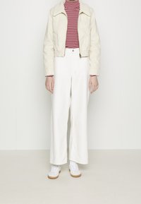 Avskuren beige jacka med fårskinnskrage över en röd och vit randig polo, kombinerad med vida, vita byxor och vita sneakers.